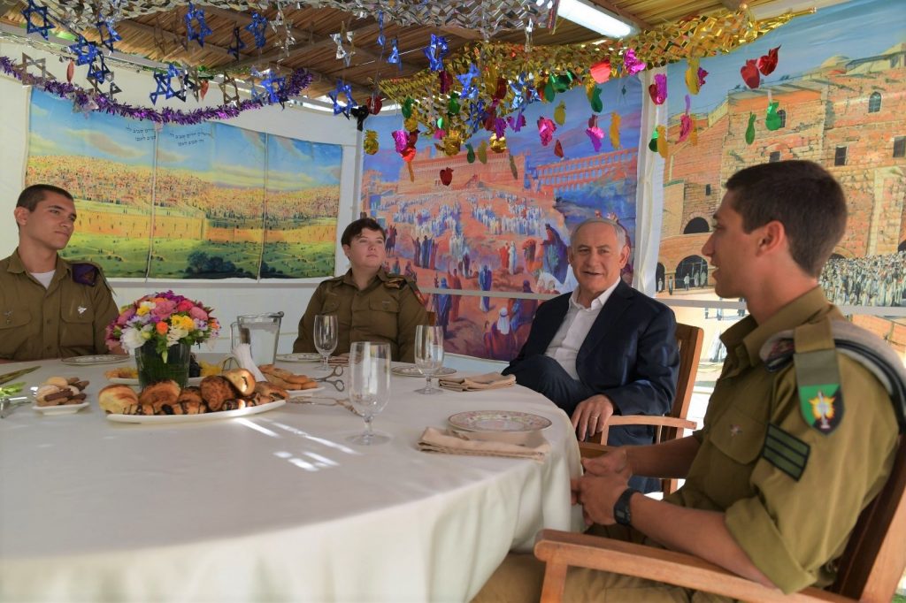 PM Netanyahu in his sukka last night with three members of the IDF. Note the decorations on the ceiling and the pictures on the walls.